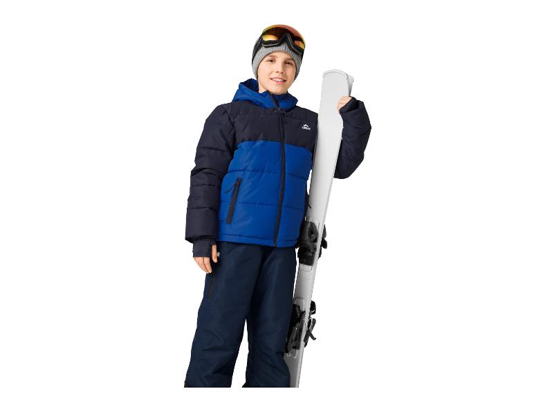 Un garçon souriant en veste de ski bleue et noire, pantalon et lunettes, tenant des skis.