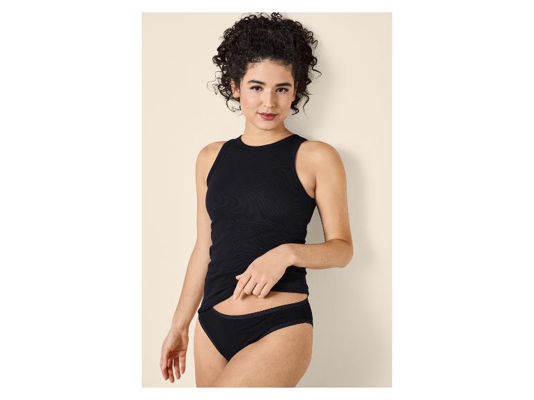 Femme aux cheveux bouclés portant un débardeur et une culotte noirs, posant sur un fond beige.