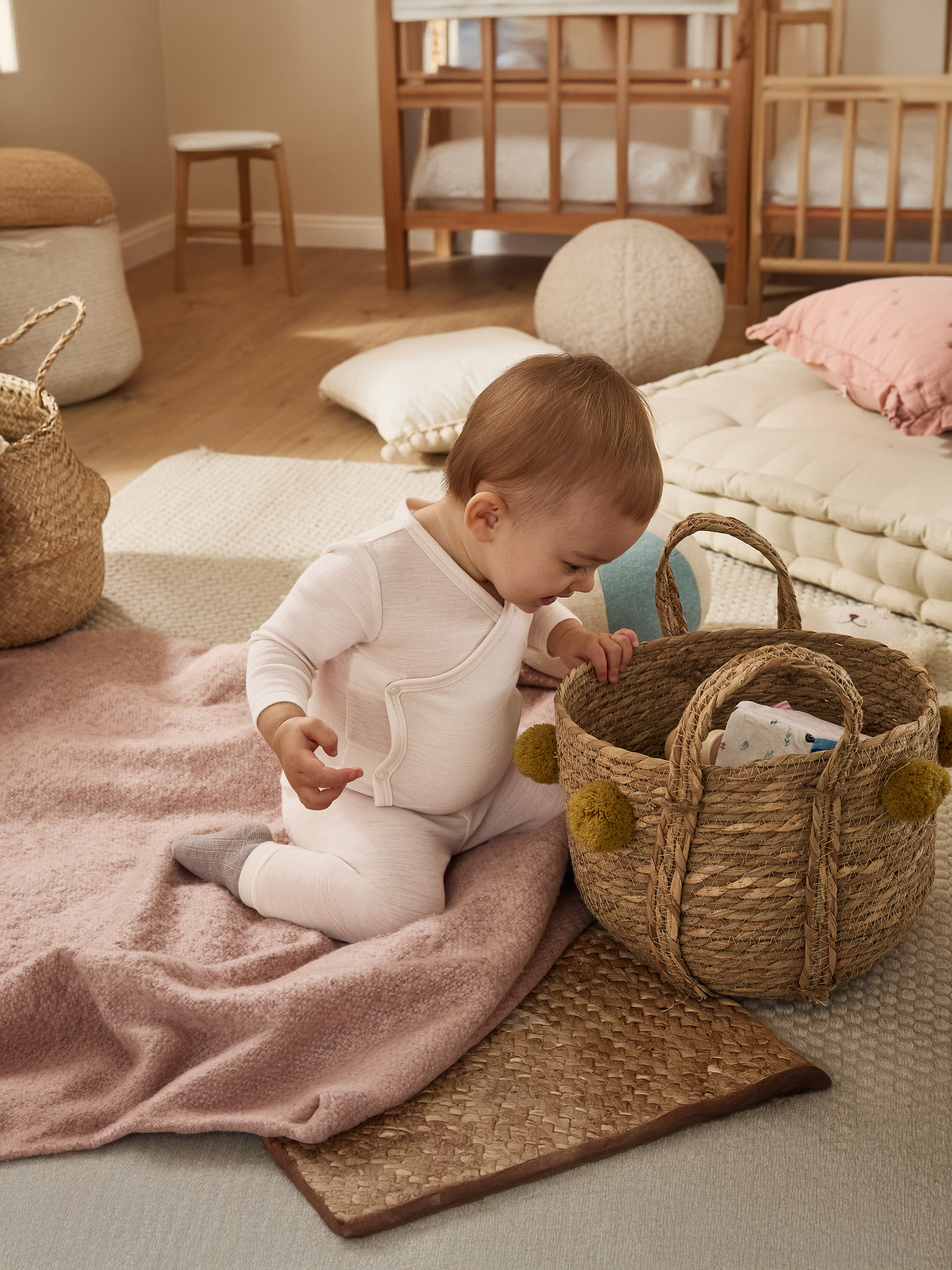 Bébé en grenouillère blanche joue avec un panier en osier sur une couverture rose.