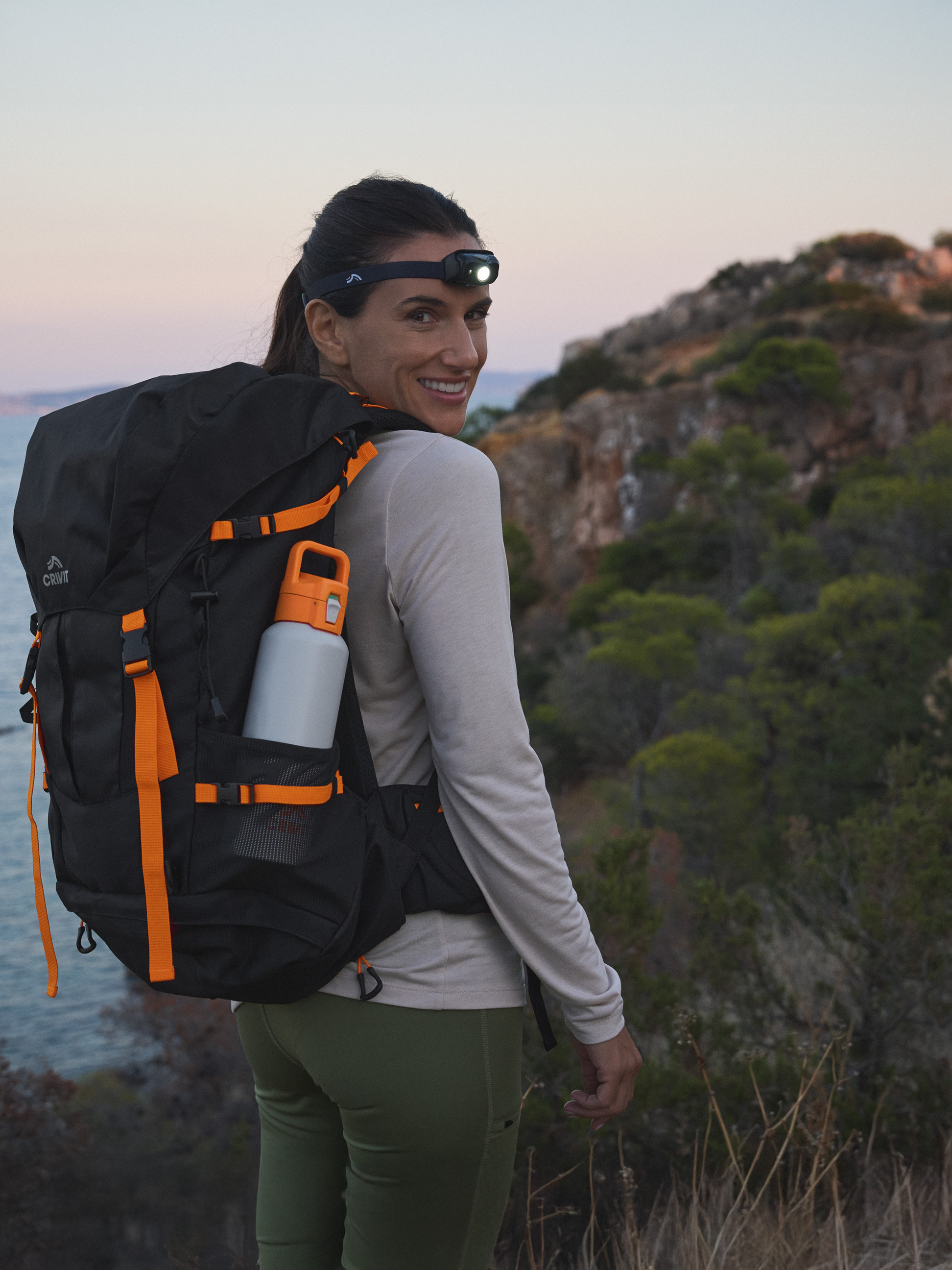 Femme avec sac à dos Crivit et lampe frontale, souriant à la caméra en pleine nature.