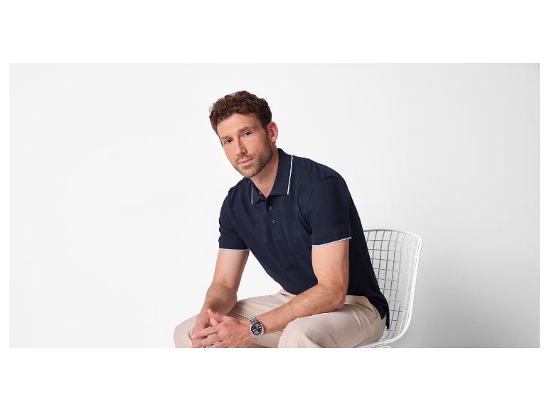 Homme en polo bleu marine et pantalon beige assis sur une chaise blanche.