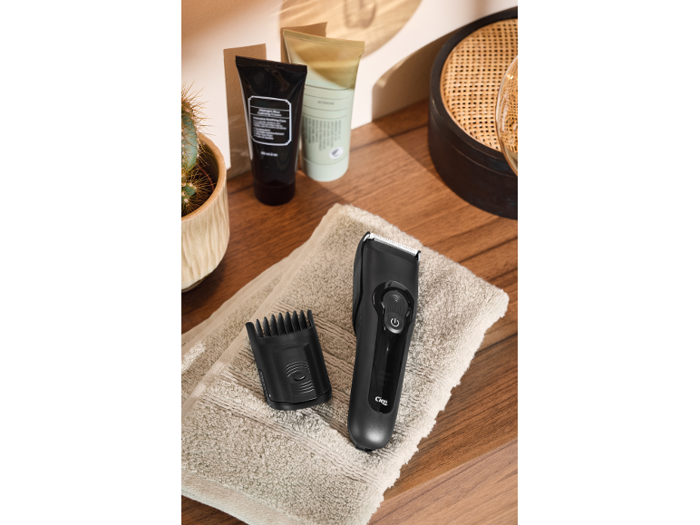Tondeuse à cheveux noire avec sabot sur une serviette, à côté de produits de soin et d'un cactus.