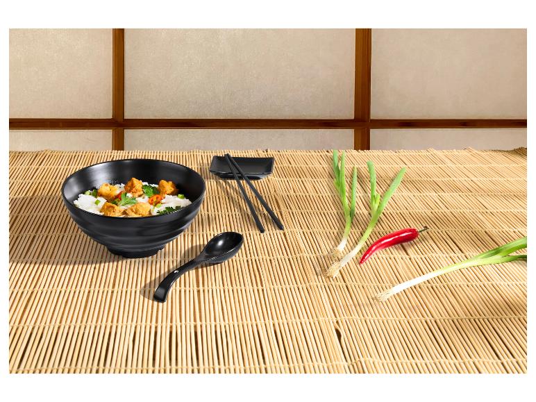 Bol noir avec de la nourriture asiatique, baguettes, cuillère, oignons verts et piment sur une natte de bambou.