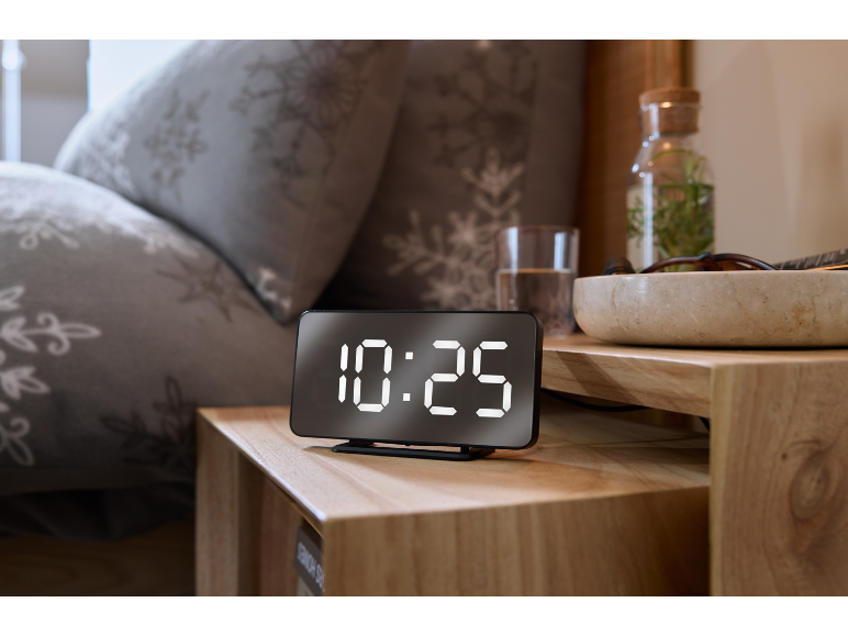 Réveil numérique affichant 10:25 sur une table de chevet en bois à côté d'un lit