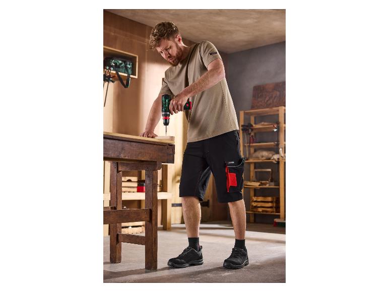 Homme en short de travail et t-shirt utilisant une perceuse sans fil sur un établi en bois.