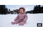 Bébé souriant en combinaison de neige rose assis dans la neige, avec une maison et des arbres en arrière-plan.