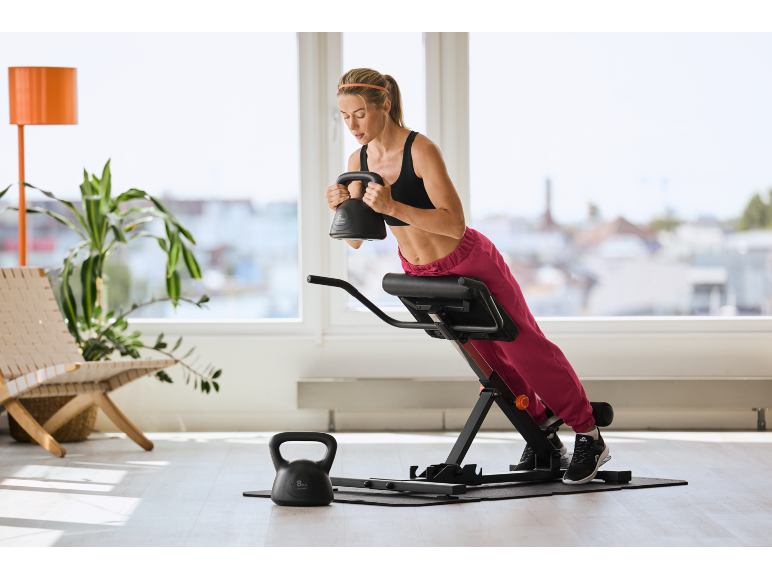 Femme s'entraînant avec un kettlebell sur un banc d'hyperextension dans une pièce lumineuse.