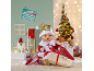 Poupée bébé en costume de Père Noël avec une hotte de cadeaux devant un décor festif.