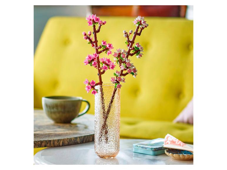 Vase avec des fleurs de cerisier Lego Botanicals sur une table, avec une tasse et une boîte en arrière-plan.