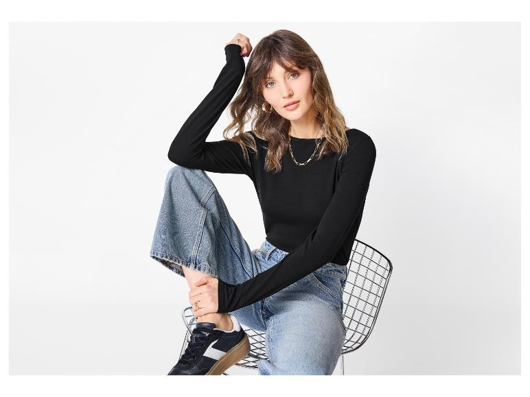 Femme en haut noir à manches longues, jean large et baskets assise sur une chaise.