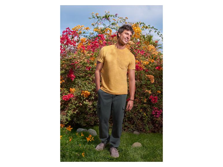 Homme en t-shirt jaune et pantalon vert foncé posant devant des fleurs.
