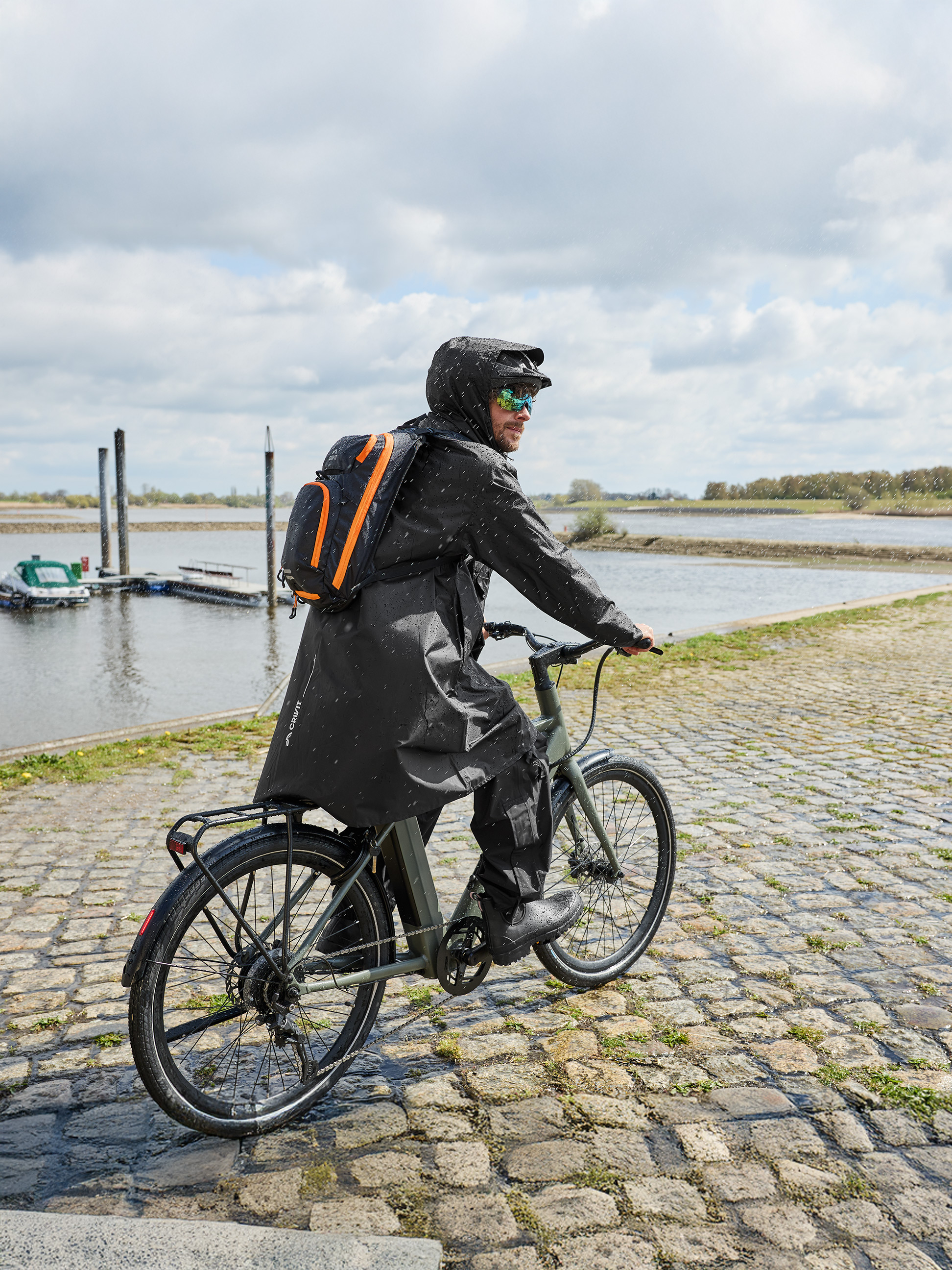 Homme en imperméable et sac à dos à vélo sous la pluie près d'une rivière.