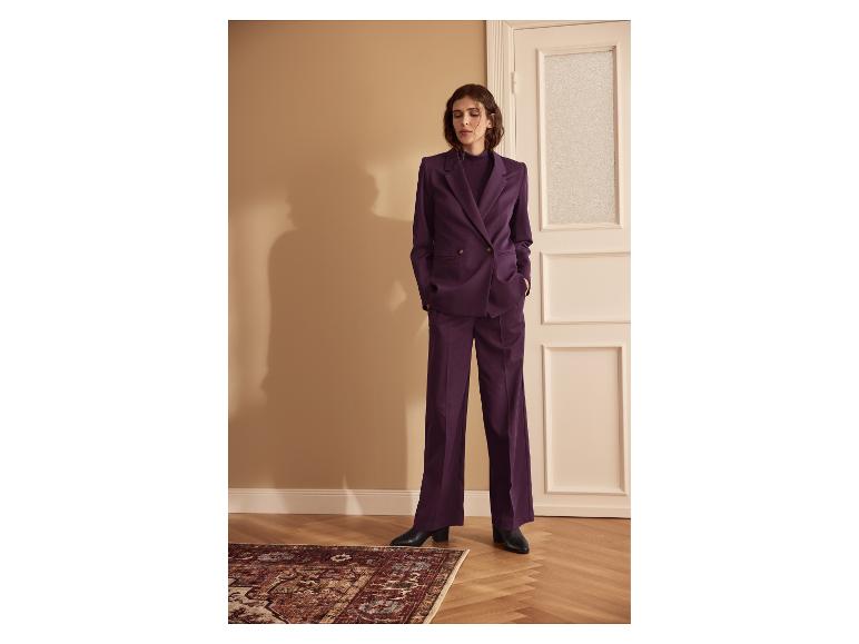 Femme en costume violet, avec blazer et pantalon, debout dans une pièce avec parquet et tapis.
