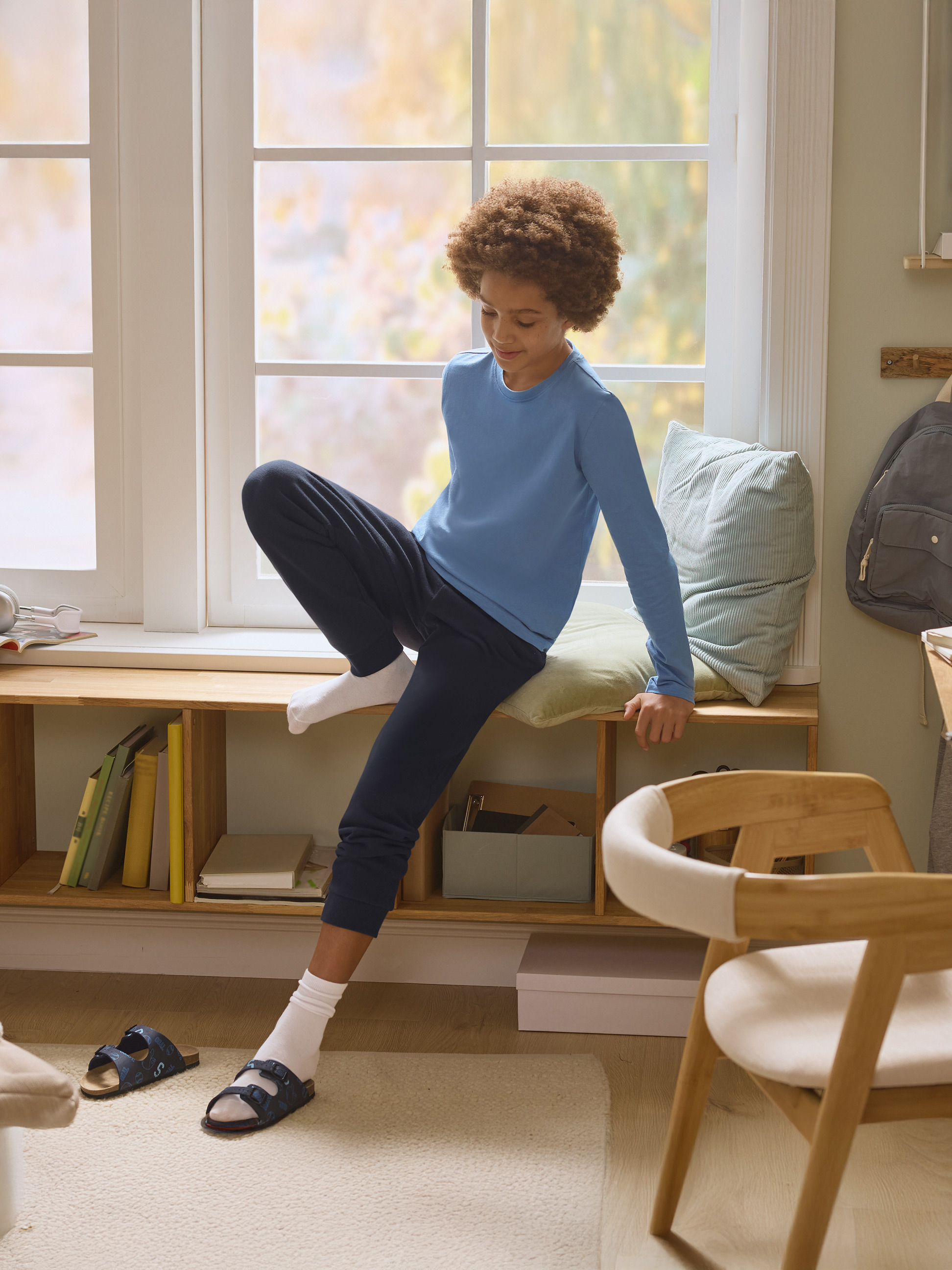 Garçon en pyjama bleu assis sur le rebord de la fenêtre avec des livres et des coussins.