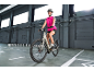 Femme souriante en tenue de sport rose et casque, à vélo électrique gris dans un entrepôt.