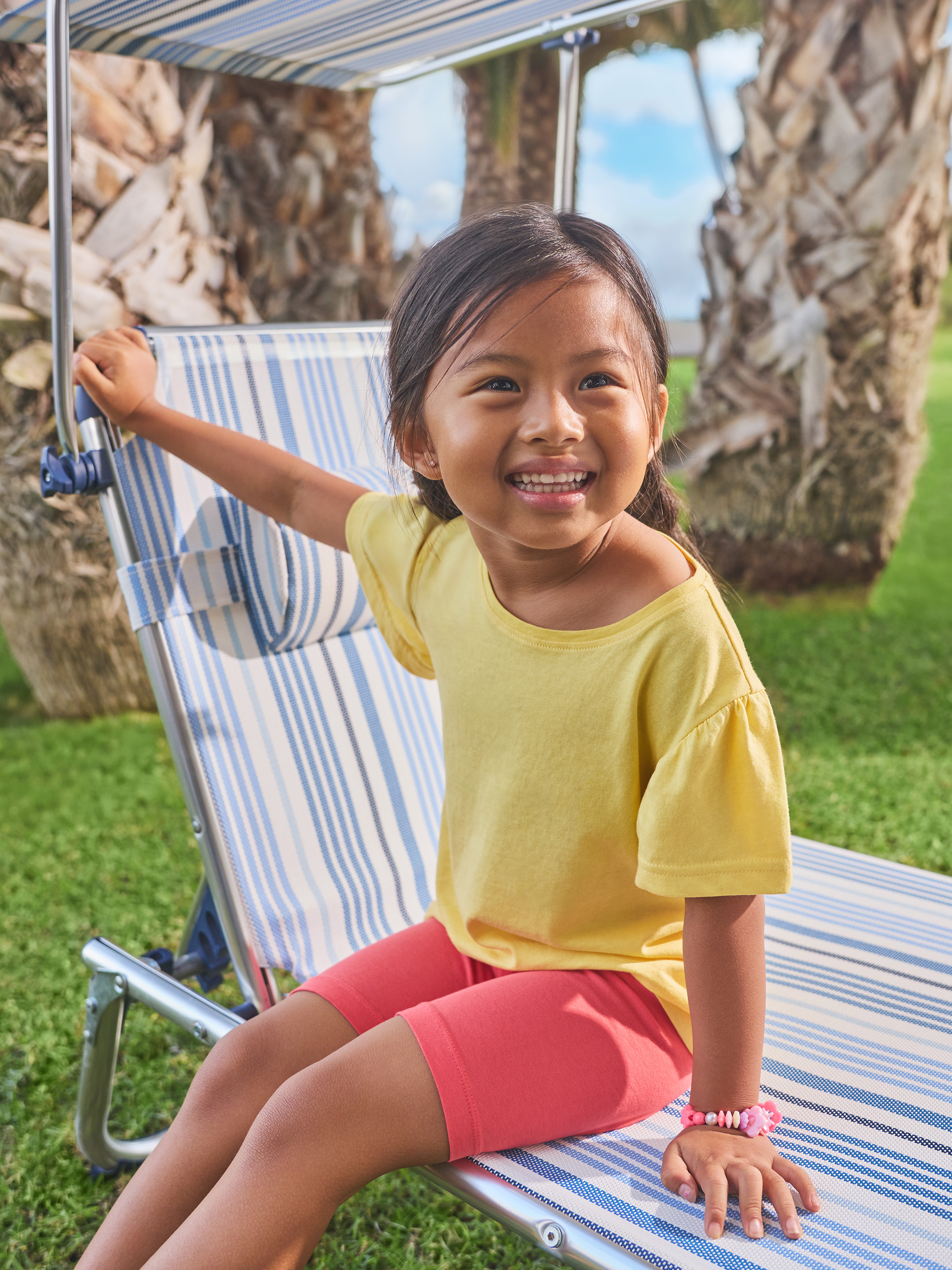 Fille souriante en t-shirt jaune et short rouge assise sur une chaise longue rayée.