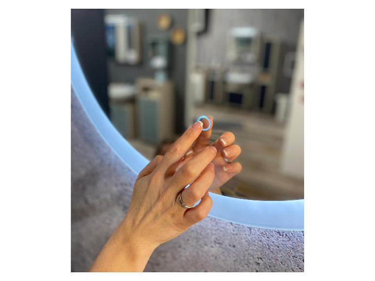 Mains féminines touchant un miroir rond éclairé, reflétant une salle de bain moderne.