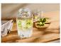 Distributeur de boissons en verre avec des citrons verts tranchés, des verres et des citrons verts entiers sur une table en bois.