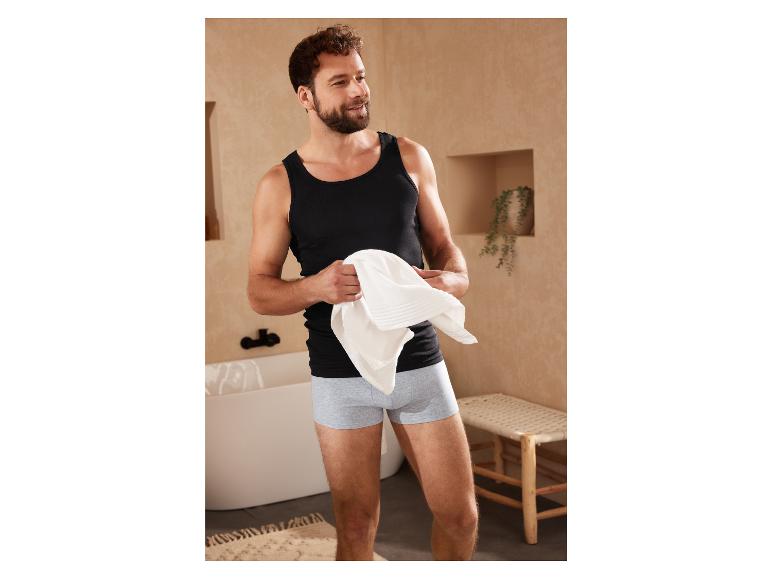 Un homme en débardeur noir et boxer gris dans une salle de bain.