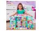 Une fille souriante tient une grande maison de poupée à plusieurs étages remplie de figurines et d'accessoires.