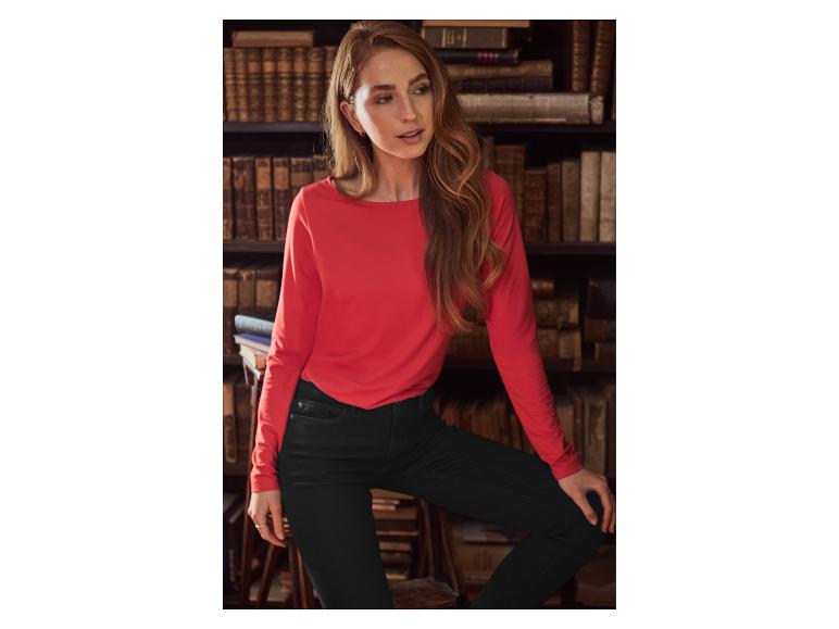 Femme en t-shirt rouge à manches longues et jean noir, appuyée sur une chaise devant une bibliothèque.