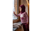 Bébé en pull rose avec ours et cagoule bordeaux regarde la neige par la fenêtre.