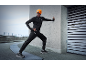 Homme en tenue de course noire et bonnet orange avec lampe frontale s'étirant contre un mur.