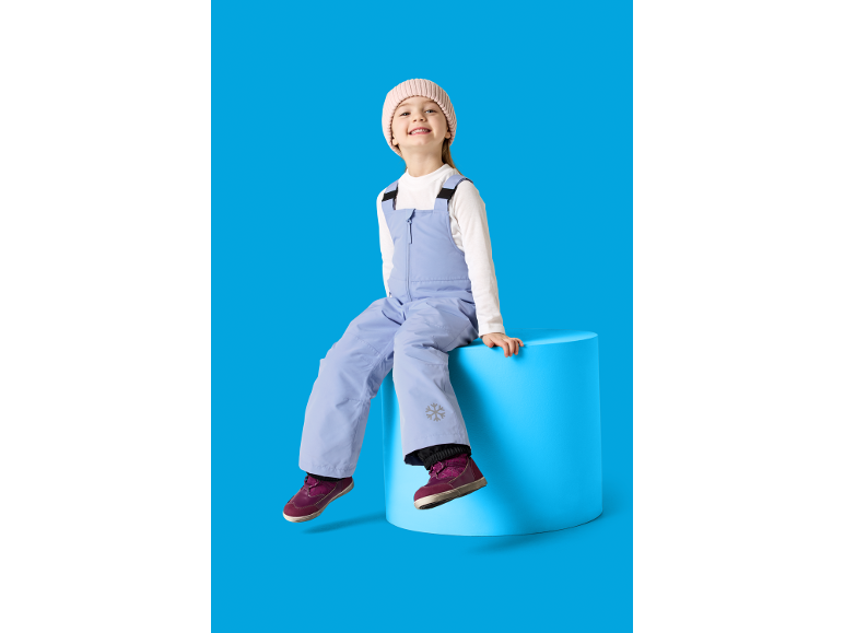 Enfant souriant en bonnet rose et combinaison de ski bleu clair avec flocon de neige, assis sur un cylindre bleu.