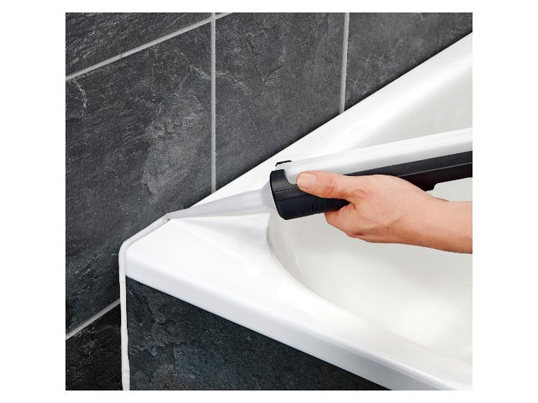 Une main applique du mastic blanc sur le bord d'une baignoire près de carreaux sombres.