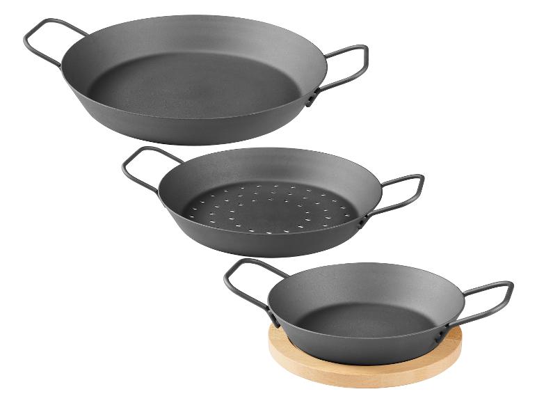 Trois poêles à paella grises avec poignées : une grande, une perforée et une petite sur un dessous de plat en bois.