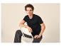 Jeune homme en t-shirt noir et jean gris foncé, assis sur une chaise blanche.