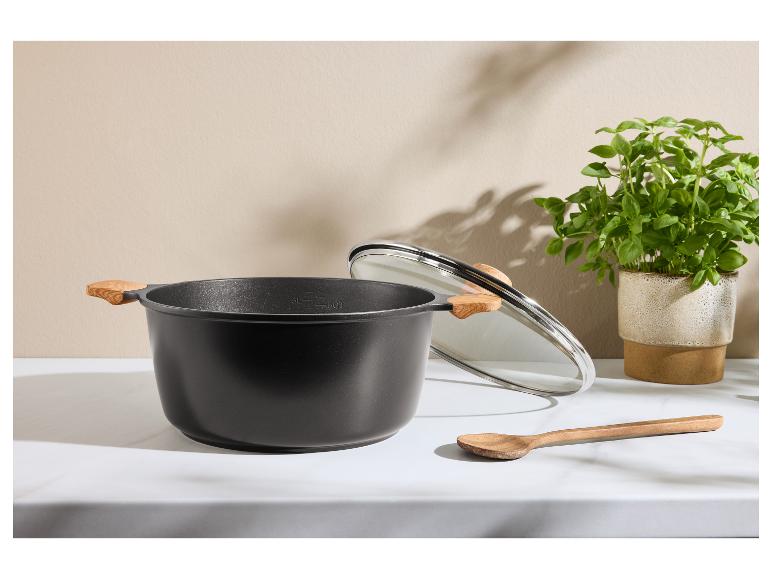 Marmite noire avec poignées en bois, couvercle en verre et cuillère en bois sur surface blanche.