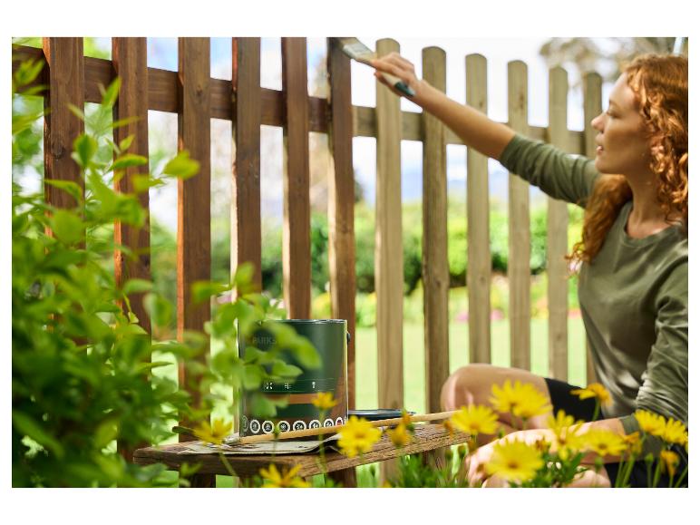 Femme peignant une clôture en bois avec de la peinture verte, un pinceau et des fleurs jaunes.