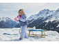 Fille en veste de ski, pantalon et gants roses, jouant avec la neige à côté d'une luge.