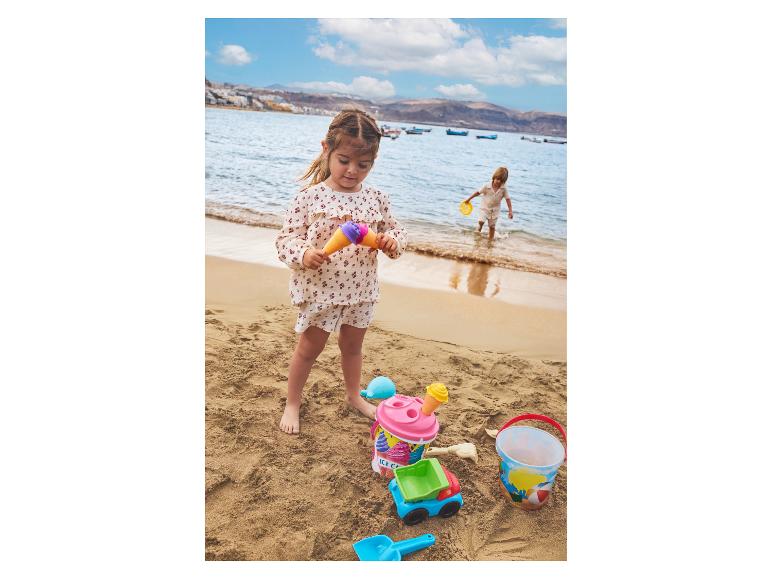 Deux enfants jouent avec des jouets de plage, dont des seaux, des pelles et un jouet en forme de cornet de glace, au bord de la mer.