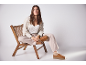 Femme assise sur une chaise en bois, porte un pantalon beige, une chemise claire et des chaussures marron.