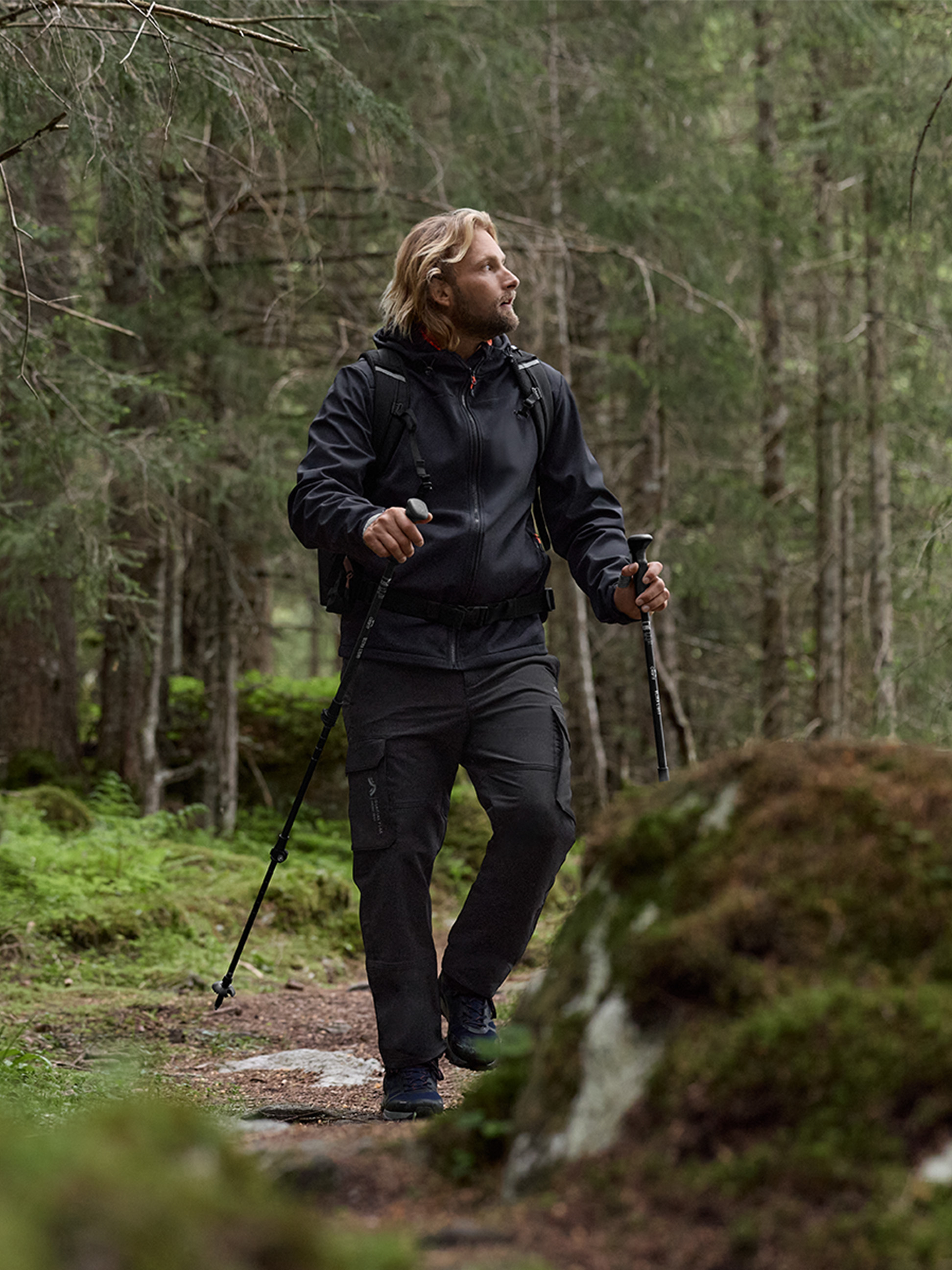 Un homme en veste de randonnée noire et pantalon cargo marche avec des bâtons de trekking en forêt.