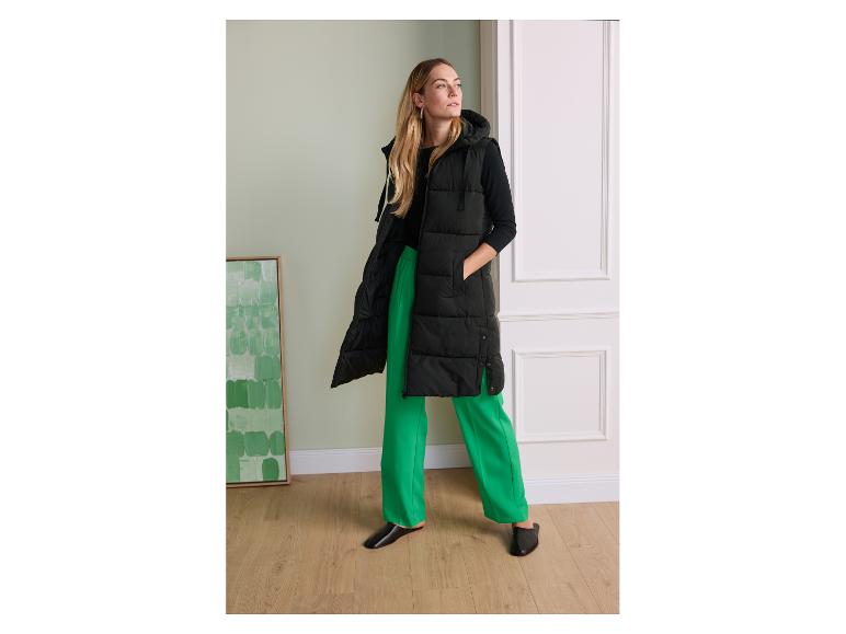 Femme en longue veste noire à capuche, chemisier noir et pantalon vert.