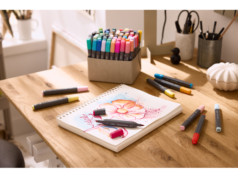 Une table en bois avec un carnet de croquis orné d'un dessin de fleur et divers marqueurs.