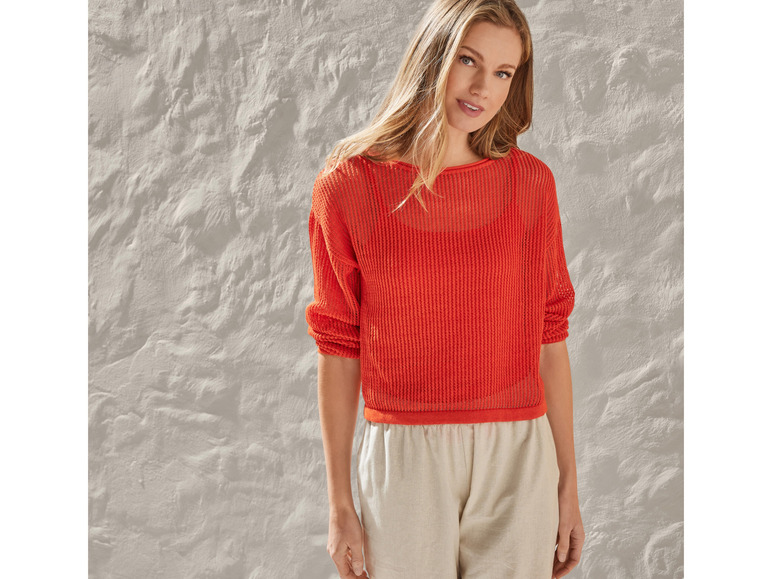 Une femme porte un pull orange en maille et un pantalon beige.
