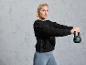 Une femme vêtue d'un sweat-shirt noir et de leggings gris tient un kettlebell noir.