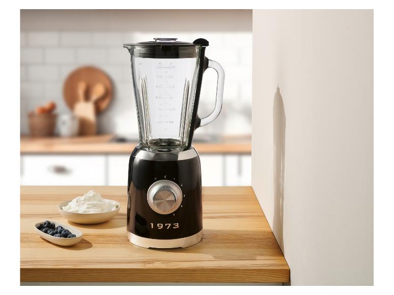 Un blender noir de style années 1973 se trouve sur un plan de travail de cuisine à côté de bols de yaourt et de myrtilles.