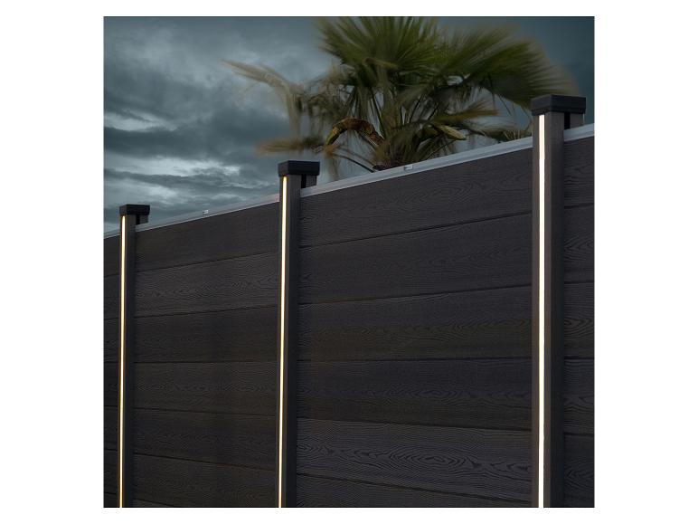 Clôture en bois foncé avec lumières LED verticales et palmier en arrière-plan.