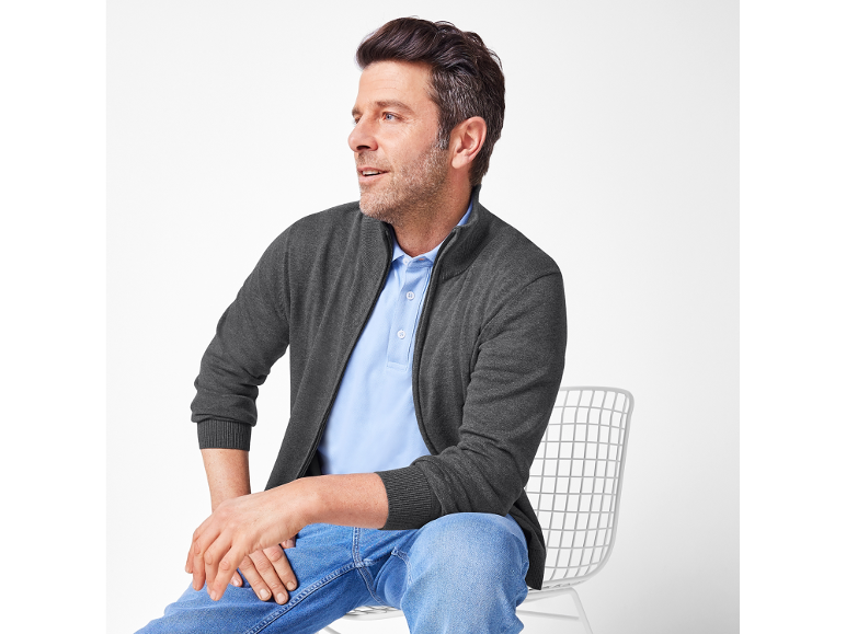 Homme en veste tricotée grise, polo bleu clair et jean, assis sur une chaise blanche.