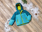 Une veste d'hiver bleue et verte pour enfants avec le texte 'Winter Fun' sur un fond en bois avec de la neige