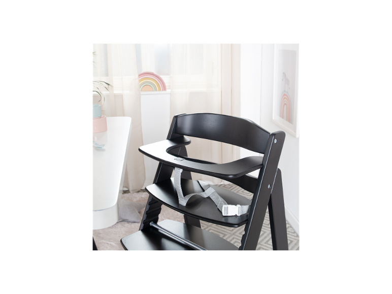 Chaise haute en bois noir avec plateau et harnais, à côté d'une table blanche avec vaisselle pour enfants.