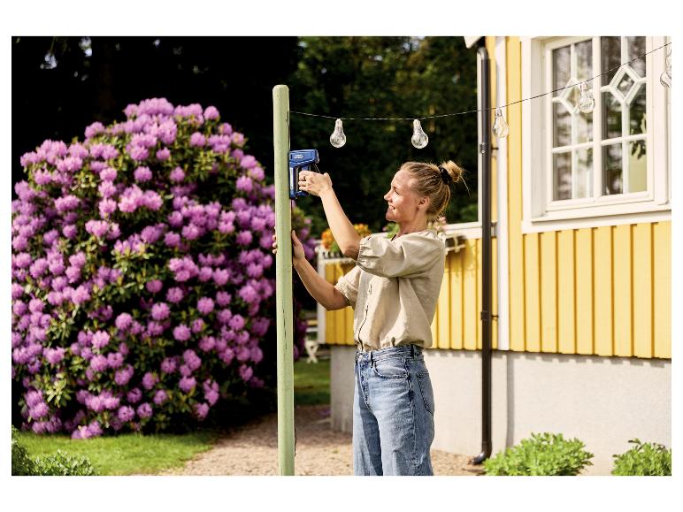 Femme utilisant l'agrafeuse manuelle LIVARNO® »RPC7« pour fixer des guirlandes lumineuses à un poteau en bois.