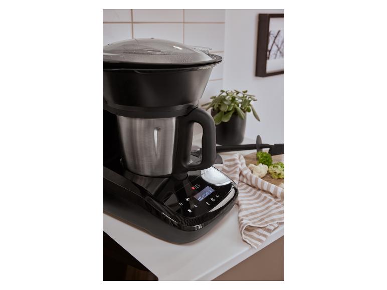 Robot de cuisine noir avec bol en acier inoxydable et couvercle transparent sur un comptoir blanc.