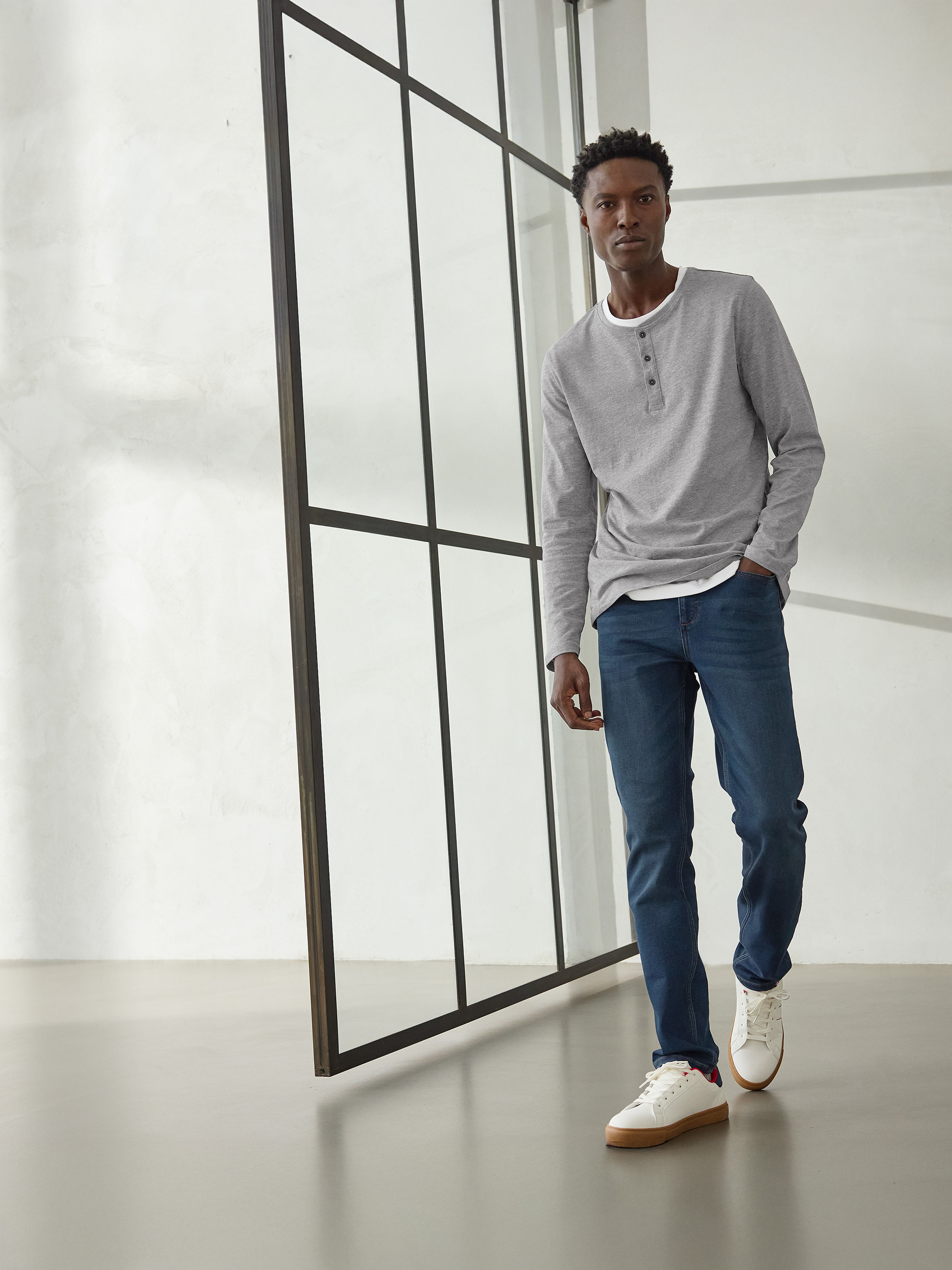 Homme en t-shirt Henley gris à manches longues, jean bleu foncé et baskets blanches.
