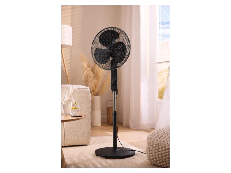 Ventilateur sur pied noir dans un salon avec canapé, lampe et fenêtre.